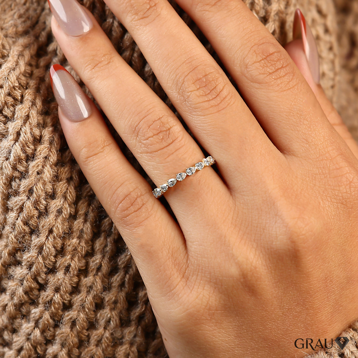 A close-up of a hand wearing a delicate silver band ring set with small clear stones, resting against a brown knit sweater. The brand name GRAU is visible in the bottom right corner.